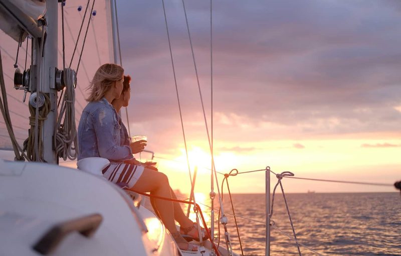 Couple of ladies are enjoying their trip on sailing boat while drinking wine and watching beautiful sunset.